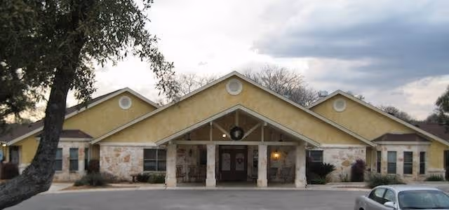 Front exterior view of a single-story building with a yellow and stone facade, a covered entrance with a wreath hanging above, and a parking area with a car in front. Trees and cloudy sky are visible in the background.