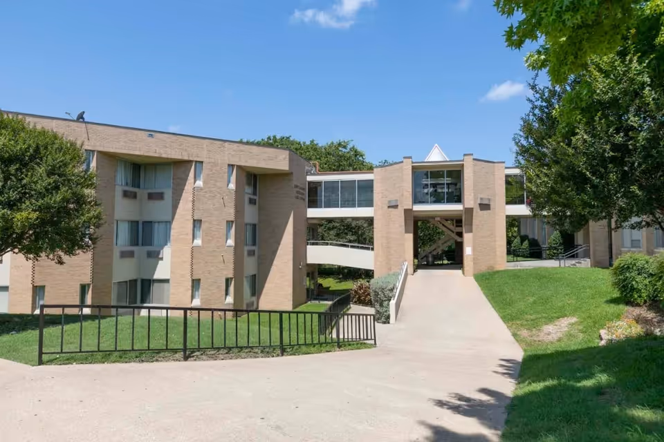 Exterior view of a multi-story brick building with large windows and a covered walkway connecting two sections. The building is surrounded by green grass, trees, and a clear blue sky.