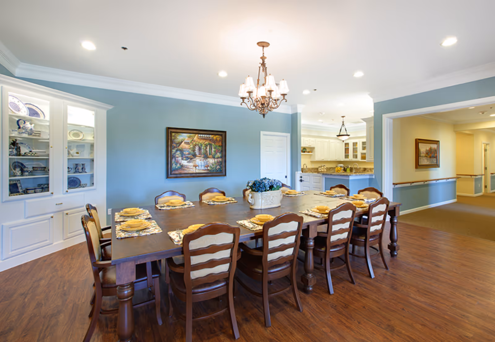 Large communal dining room with a long wooden table set for place settings, chandelier, display cabinet, and an open view into a kitchen and hallway.