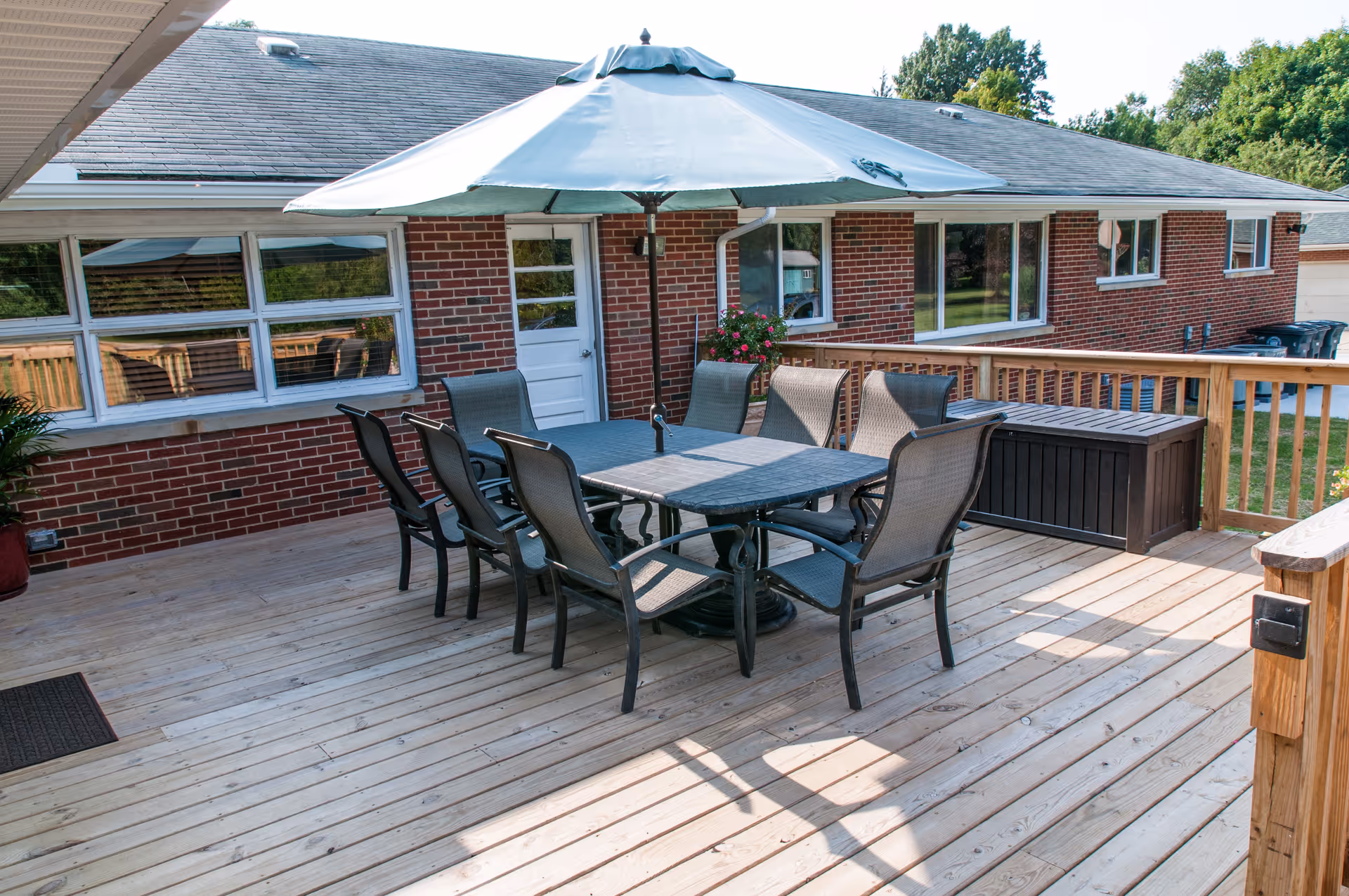 Wooden deck patio with a dining table, umbrella and several chairs next to a brick building.