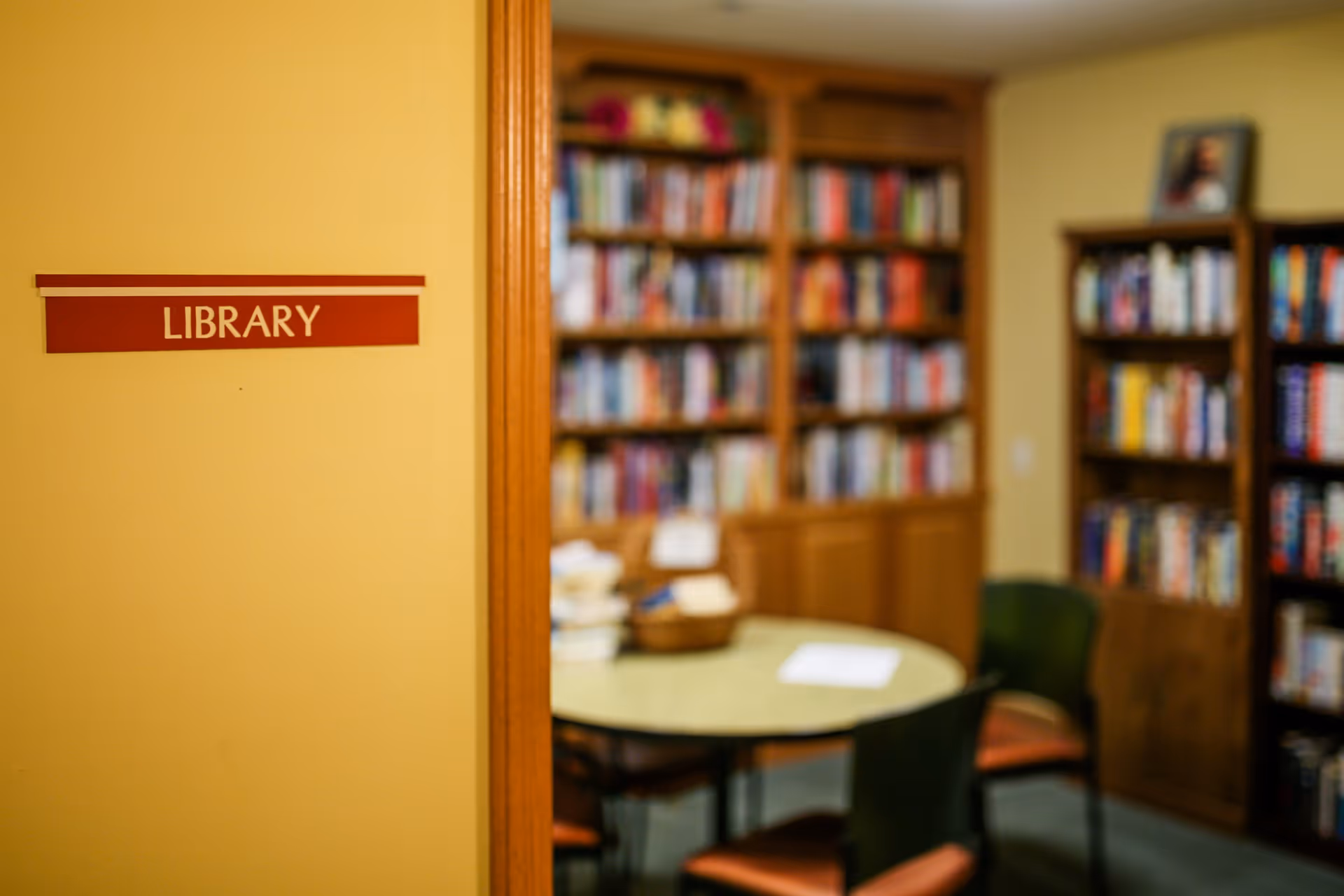 Interior view of a library room with wooden bookshelves filled with books, a round table with chairs around it, and a sign on the wall outside the room that reads 'LIBRARY'.