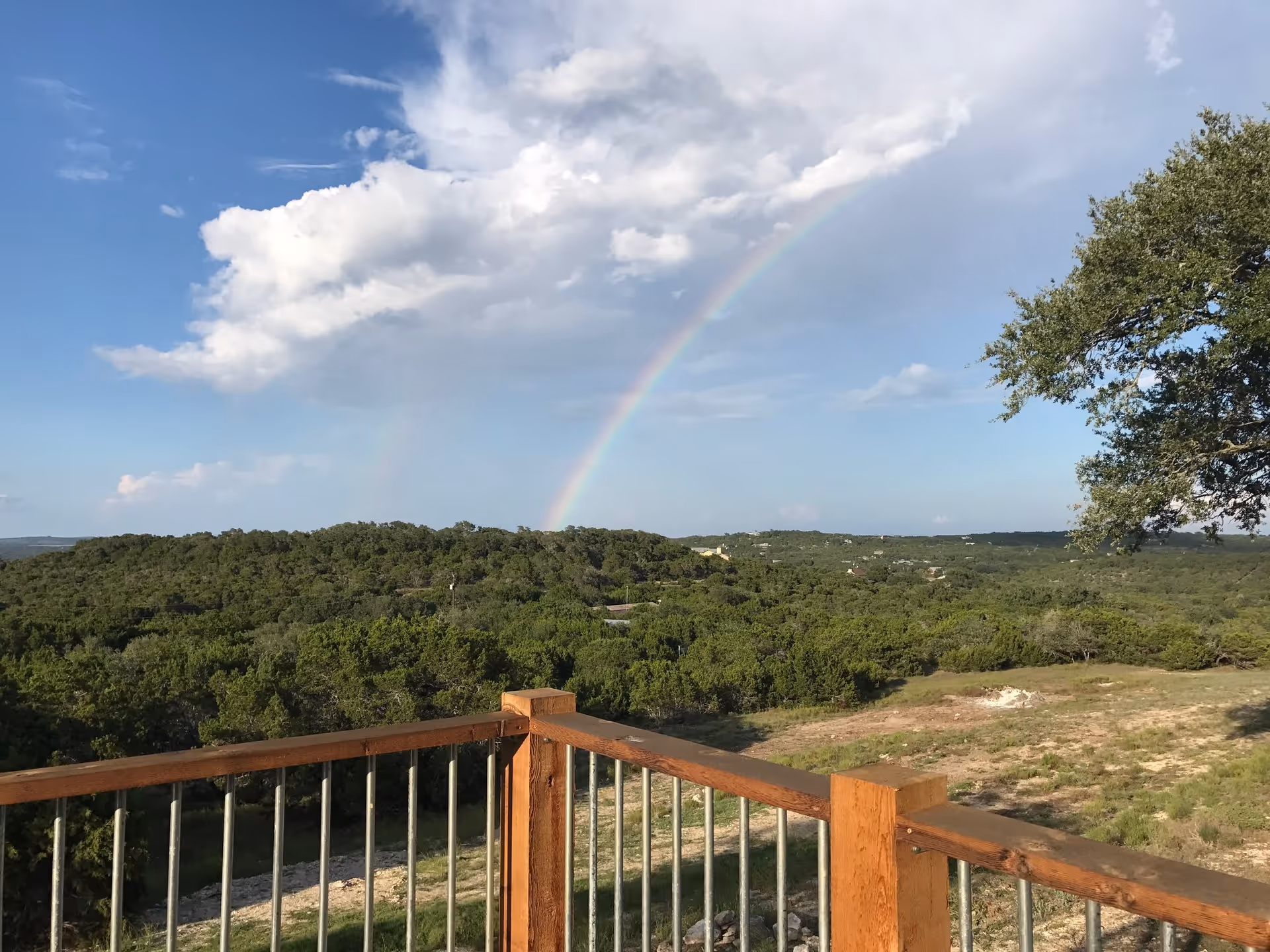View from a wooden deck overlooking green hills and trees with a rainbow arcing across a partly cloudy sky.