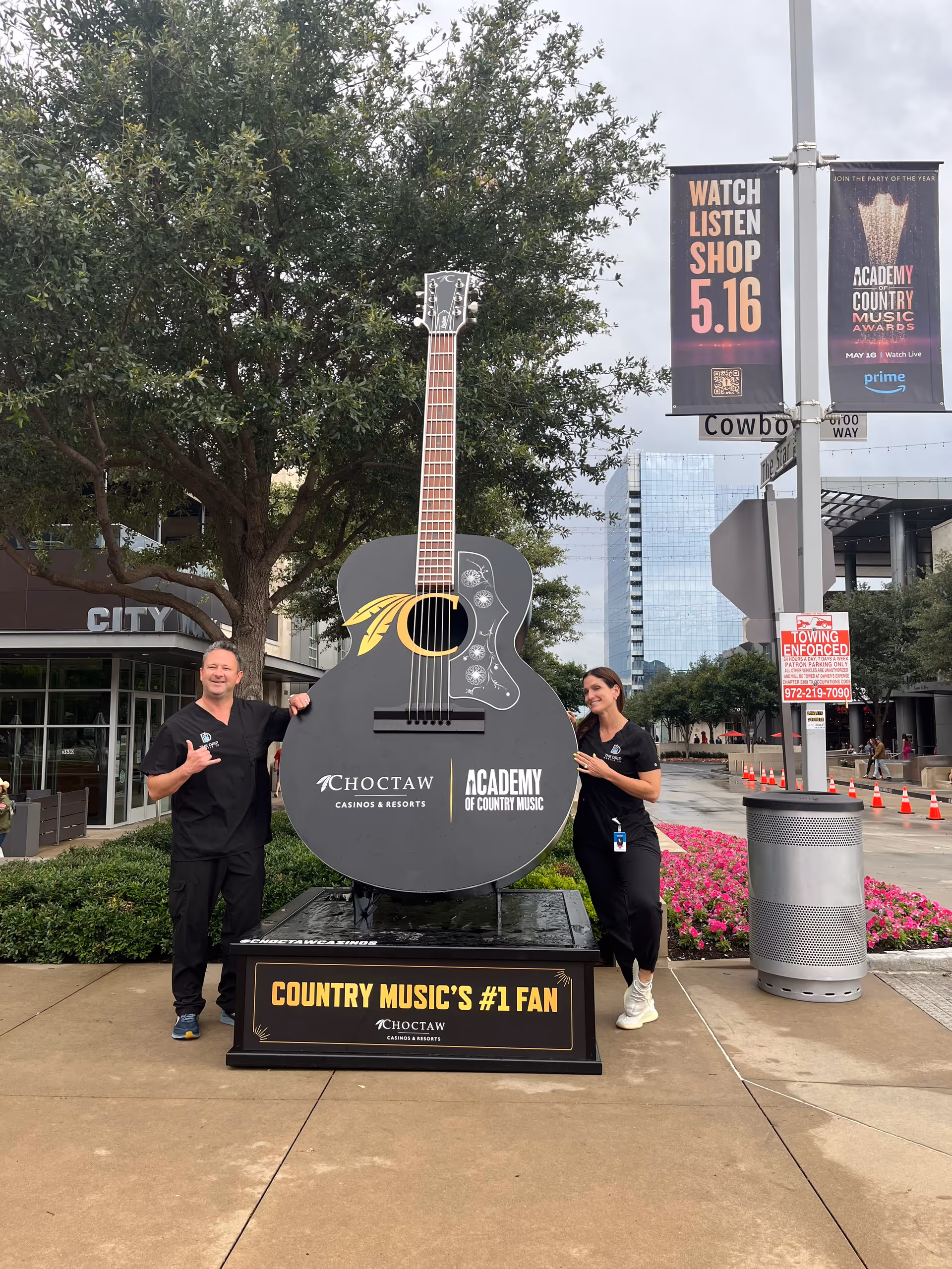 Two staff members pose beside a large guitar sculpture outdoors near urban buildings and banners.