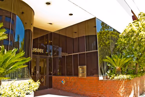 Glass-front building entrance with brick base, planters, and surrounding greenery.