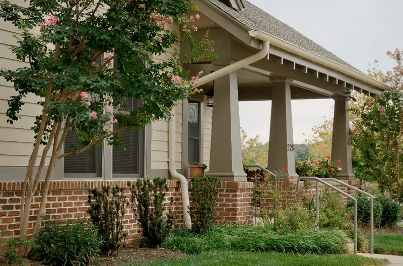 Exterior view of a residential building with a covered porch supported by large columns, brick foundation, and beige siding. There are flowering trees and shrubs in the landscaped yard, and a metal handrail along the steps leading to the porch.