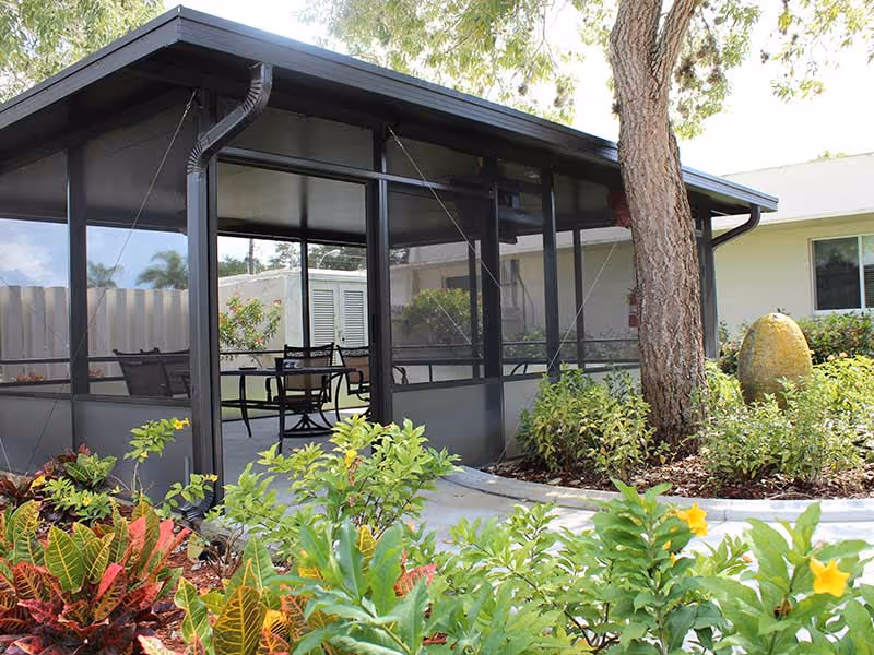 A screened-in outdoor patio area with a table and chairs, surrounded by lush green plants and colorful foliage. A large tree and a yellow decorative stone are visible near the patio, with a building in the background.