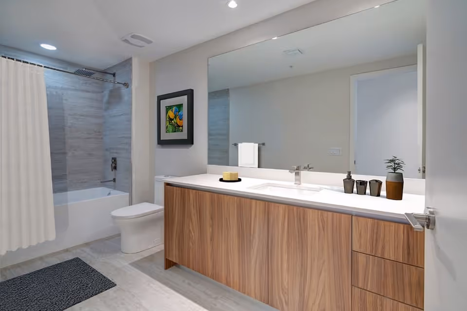 Modern bathroom with a large mirror above a wooden vanity with a white countertop. The vanity has a sink with a faucet, a small potted plant, and decorative items. To the left, there is a white toilet and a bathtub with a shower curtain. The walls and floor have light-colored tiles, and a framed picture hangs on the wall above the toilet.