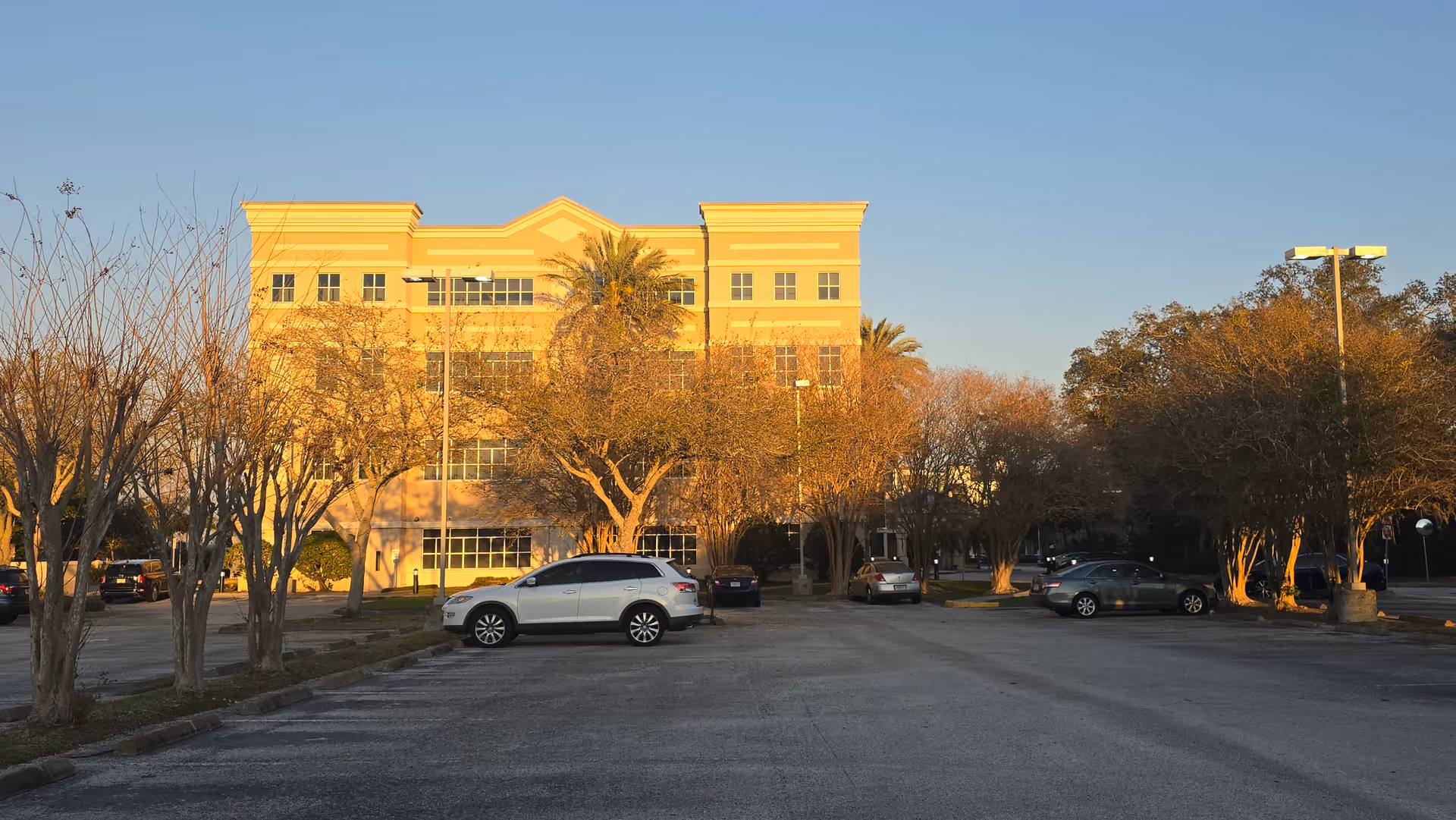 Exterior of a senior living facility with a parking lot and trees in warm sunset light.