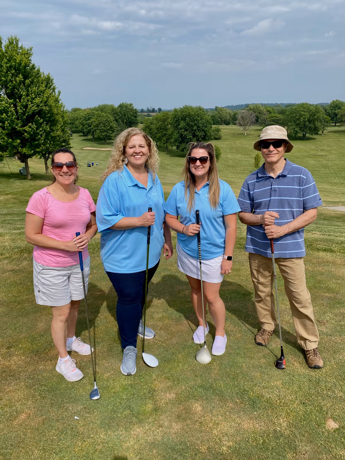 Four people pose with golf clubs on a sunny, grassy outdoor course.