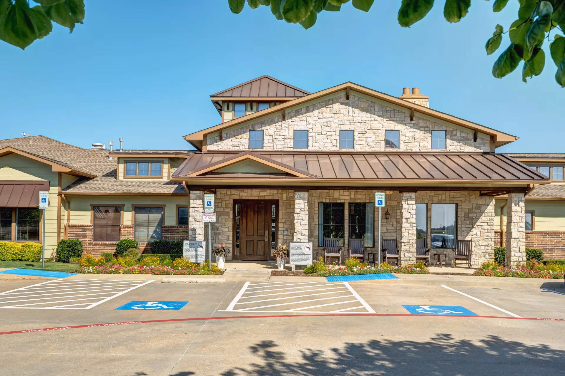 Front exterior view of Buffalo Creek Assisted Living and Memory Care facility with stone and brick facade, a covered entrance with wooden chairs, and designated handicapped parking spaces in front.