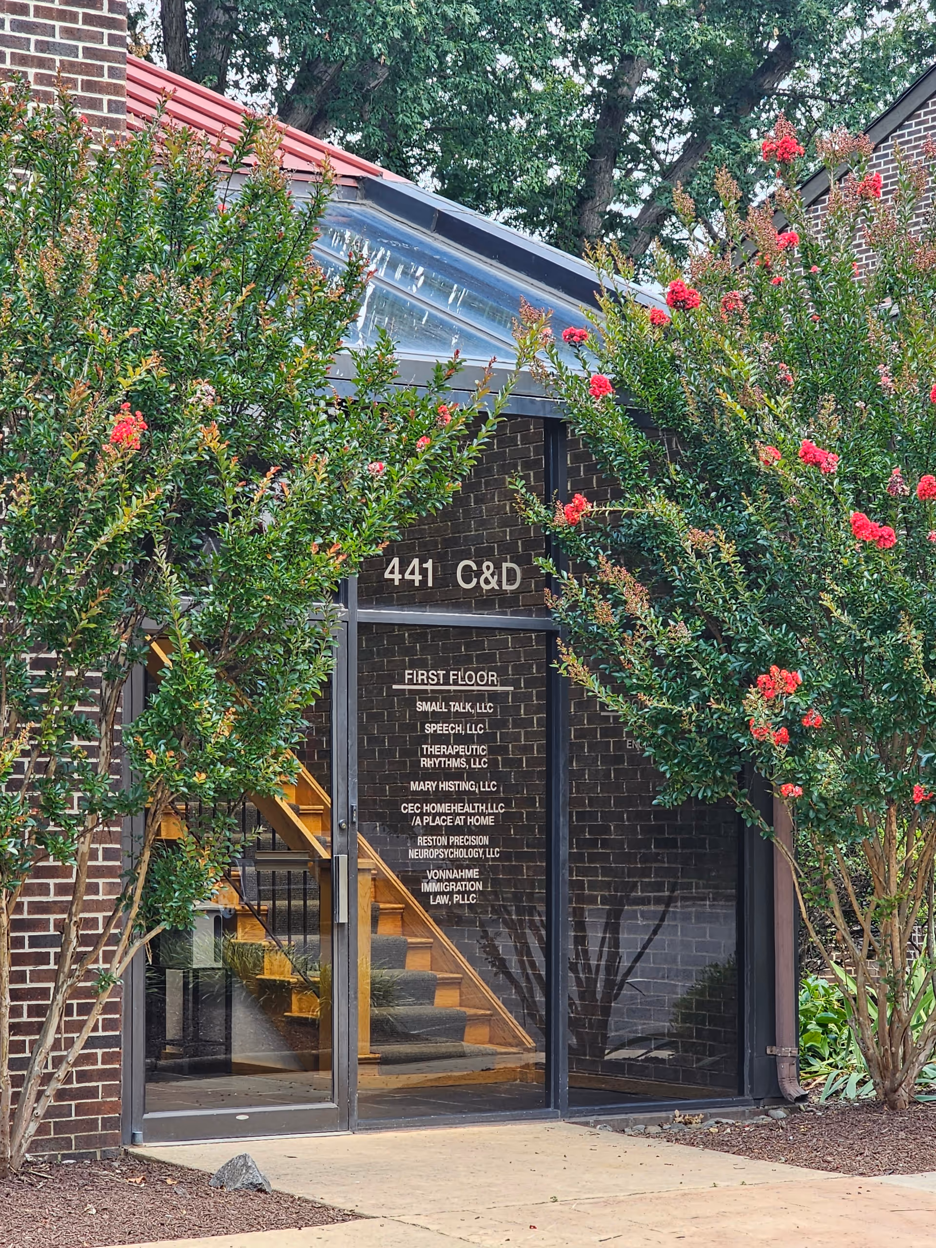 Brick building entrance with glass doors, stairs visible inside, and flowering trees framing the doorway.