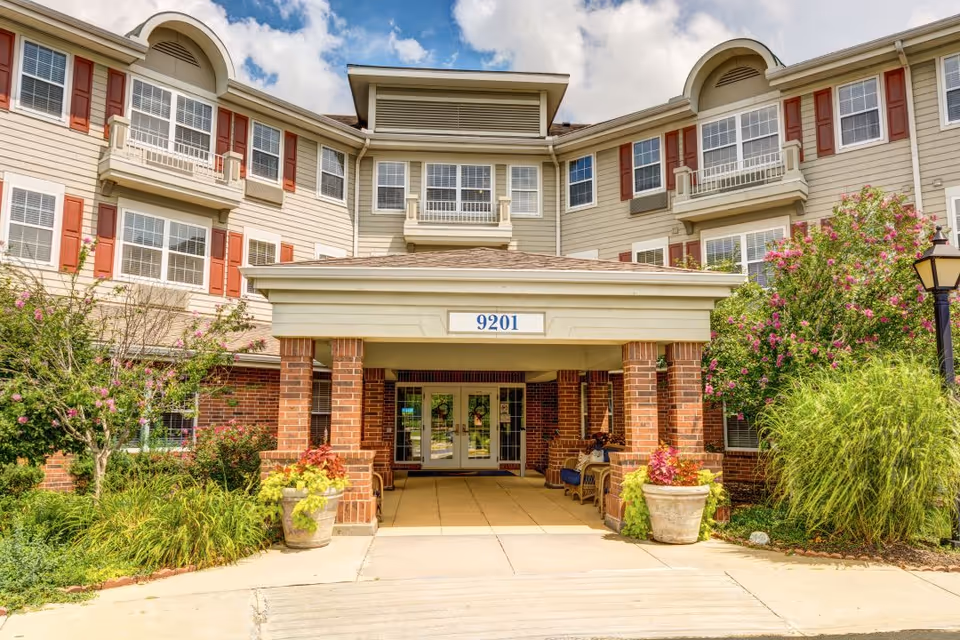 Front entrance of a multi-story senior living facility with beige siding, red shutters, and a covered entryway supported by brick columns. The building number 9201 is displayed above the entrance. There are landscaped bushes, flowering plants, and a lamppost near the walkway leading to the doors.