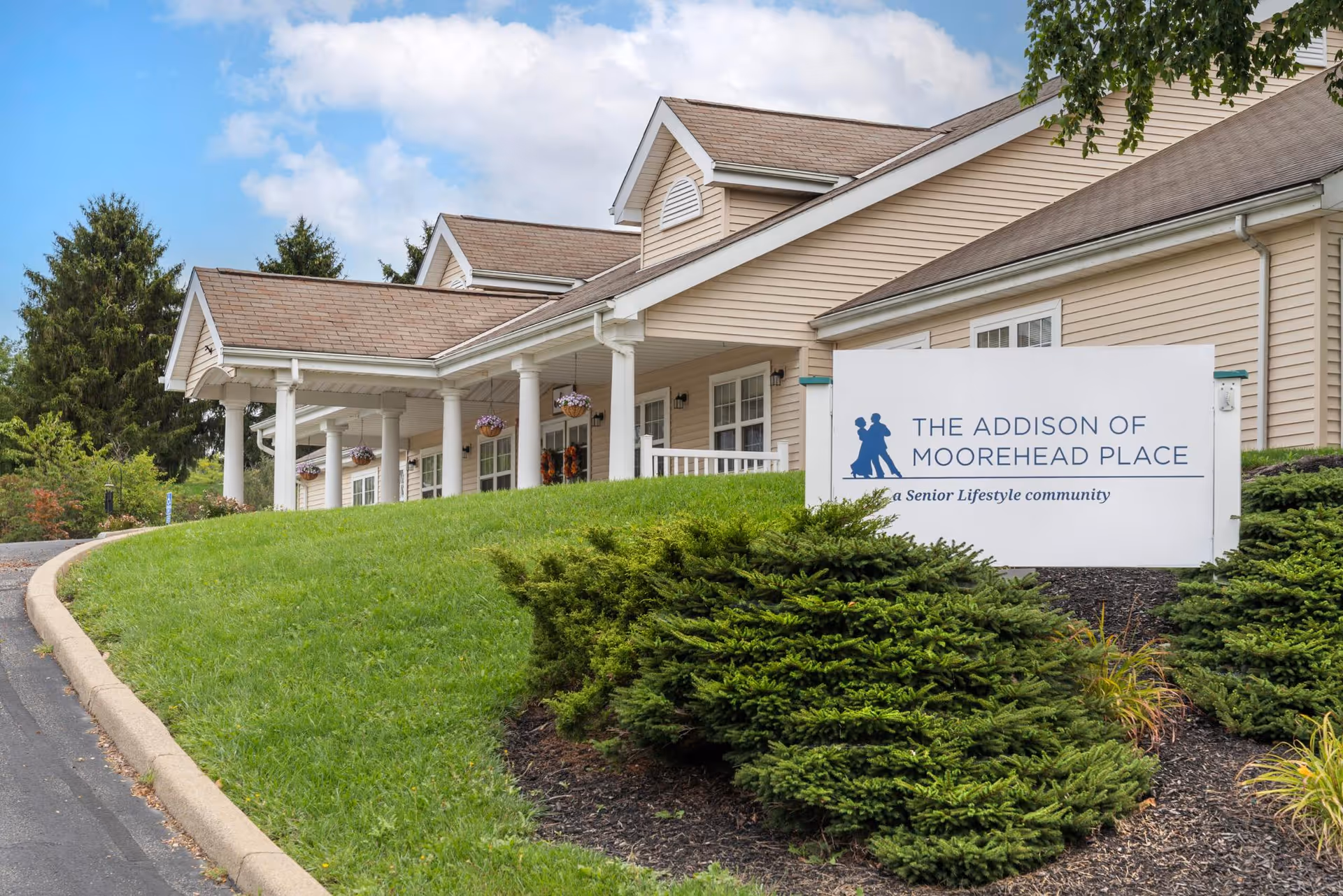 Exterior view of The Addison of Moorehead Place senior living community building with beige siding, white columns, and a sloped roof. There is a green lawn and shrubs in front, along with a white sign displaying the facility's name and logo.