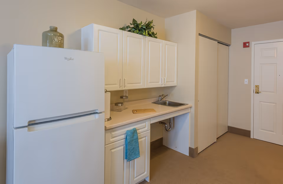White kitchen area with refrigerator, cabinets, sink, and towel in a senior living facility.
