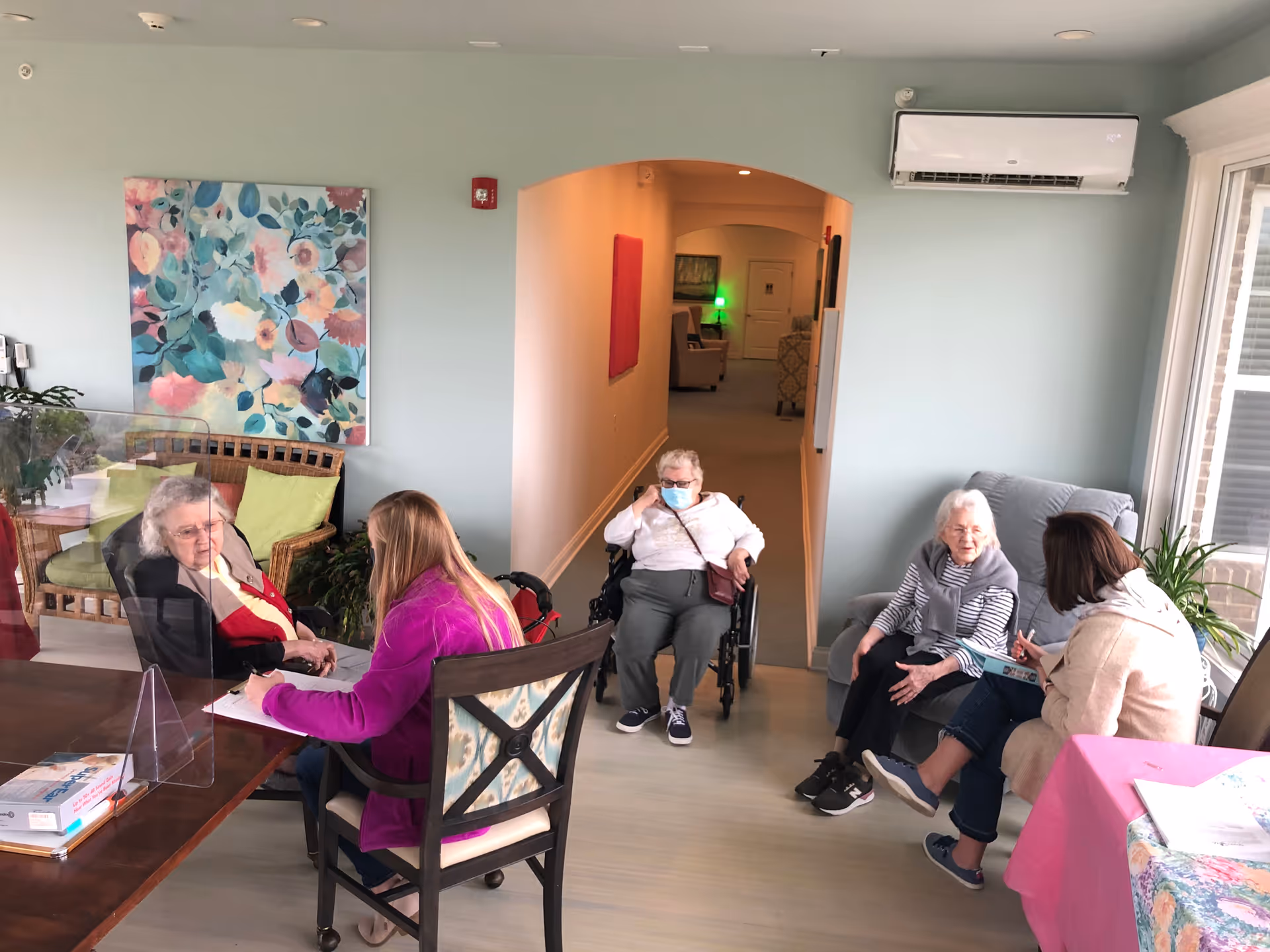 A group of elderly residents and staff talking in a bright assisted living common room, with one resident in a wheelchair.
