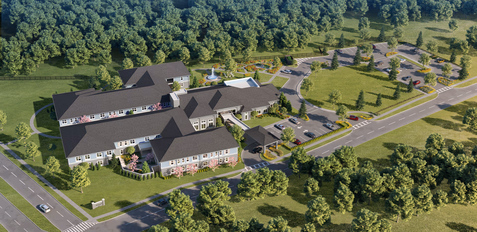 Aerial view of HearthStone at Nona Lakes senior living facility surrounded by trees and greenery. The building has multiple wings with dark roofs and light-colored walls. There is a parking lot with several cars, landscaped gardens, and a fountain visible on the grounds. Roads and sidewalks lead to the facility entrance.