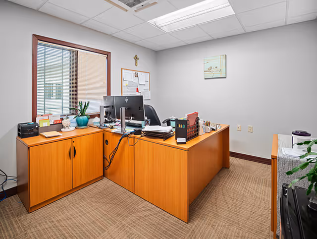 Office desk area with computer, filing trays, and bulletin board in a neutral room.