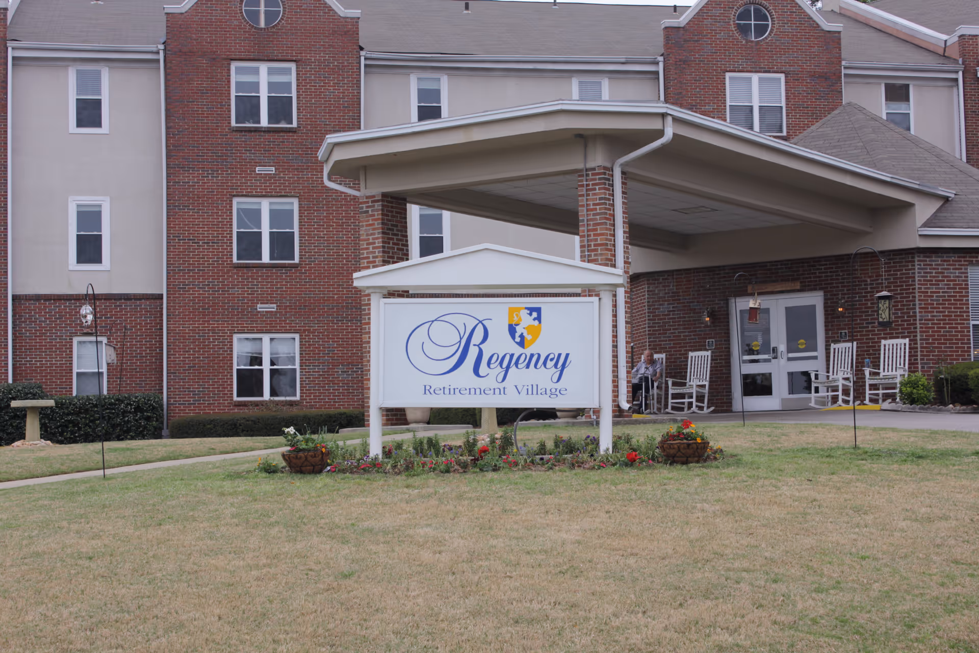 Exterior view of Regency Retirement Village of Birmingham showing a brick building with multiple windows and a covered entrance. In front of the entrance is a white sign with the facility's name and logo, surrounded by a small flower bed. There are white rocking chairs on the porch and a person sitting near the entrance.