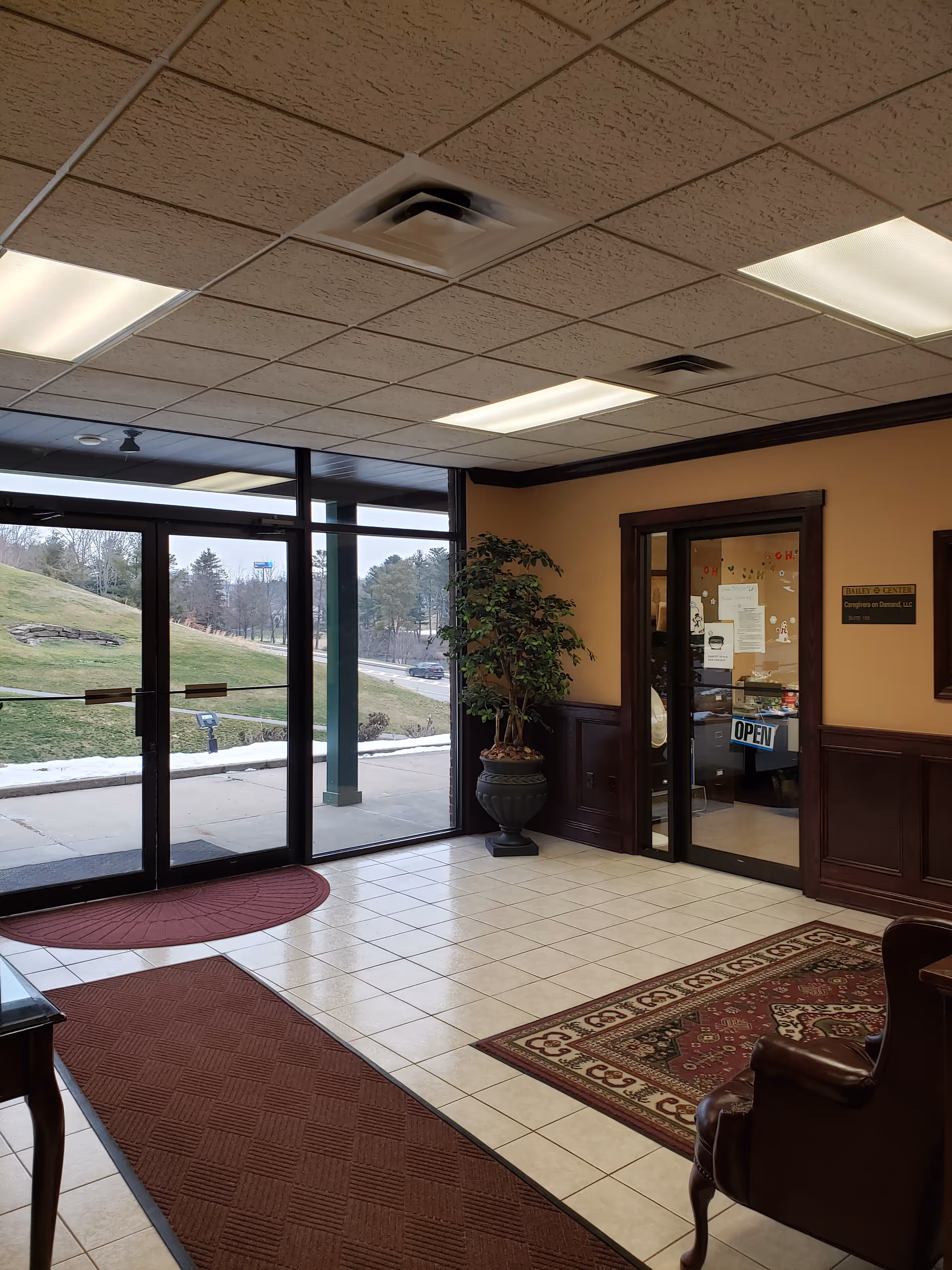 Interior senior living lobby with glass doors, tiled floor, potted plant, and rug.