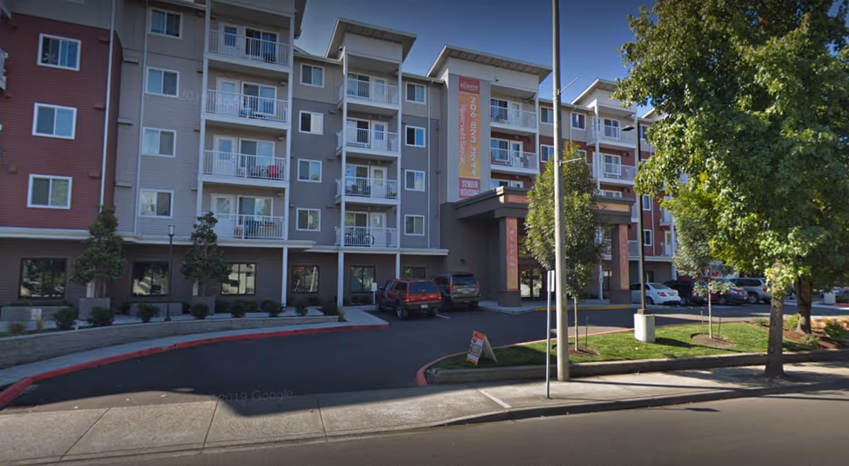 Multi-story senior living building with balconies and a vehicle-filled driveway at the main entrance.
