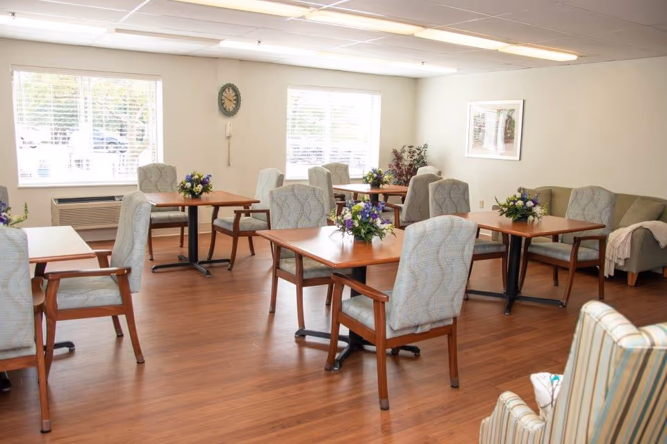 A bright senior living dining room with several wooden tables, upholstered chairs, and floral centerpieces.