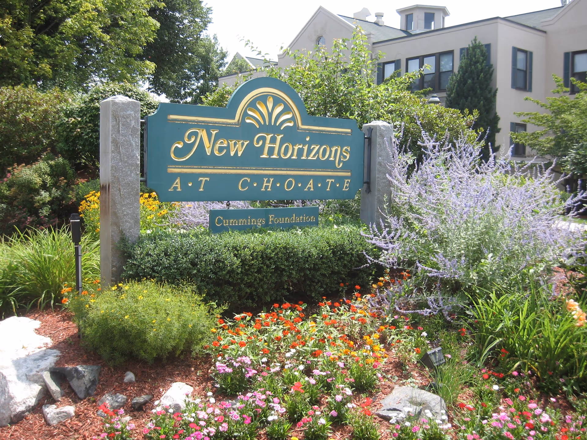 A green and gold sign reading 'New Horizons At Choate Cummings Foundation' is surrounded by colorful flowers and greenery, with a large building visible in the background under a clear sky.