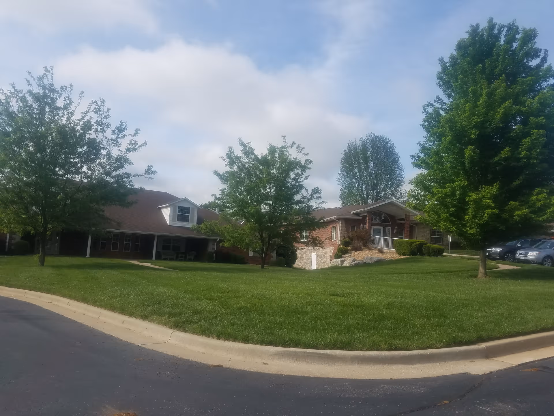 Single-story brick senior living facility with a green lawn, trees, and parked cars nearby.