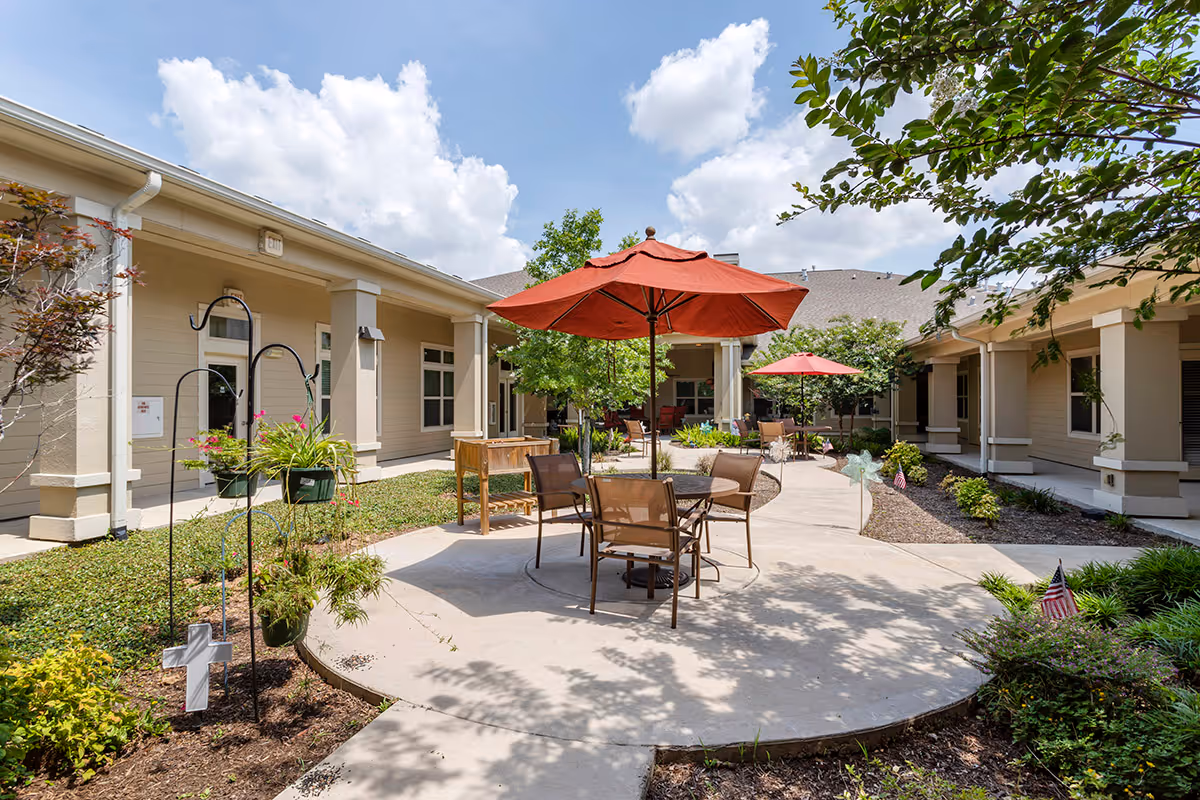Outdoor courtyard area at Caydance Assisted Living & Memory Care featuring a paved walkway, patio tables with red umbrellas, chairs, and surrounding greenery under a partly cloudy sky.