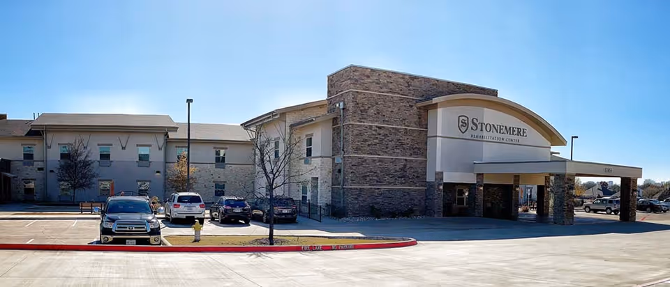 Front exterior of the Stonemere Rehabilitation Center building with a covered entrance, parking lot, and several cars under a clear blue sky.