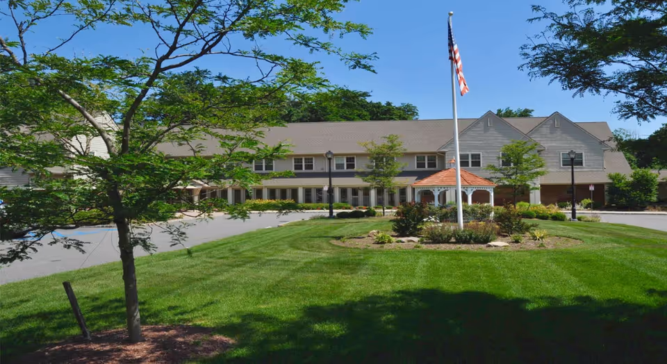 Front view of a two-story senior living facility with an American flag, small gazebo, and landscaped lawn and trees.