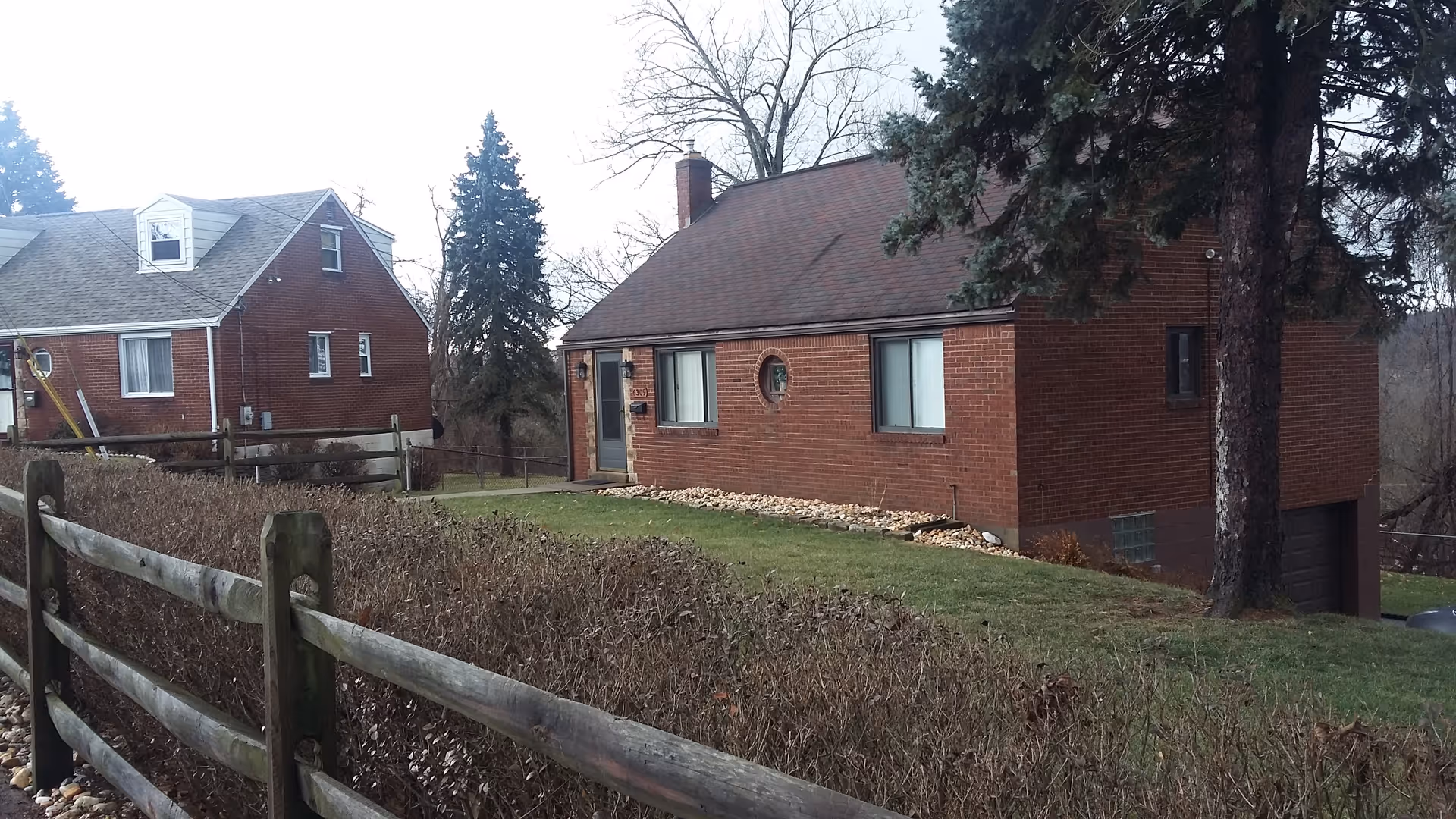 Two brick houses on a grassy hillside, with a wooden fence and tall trees.