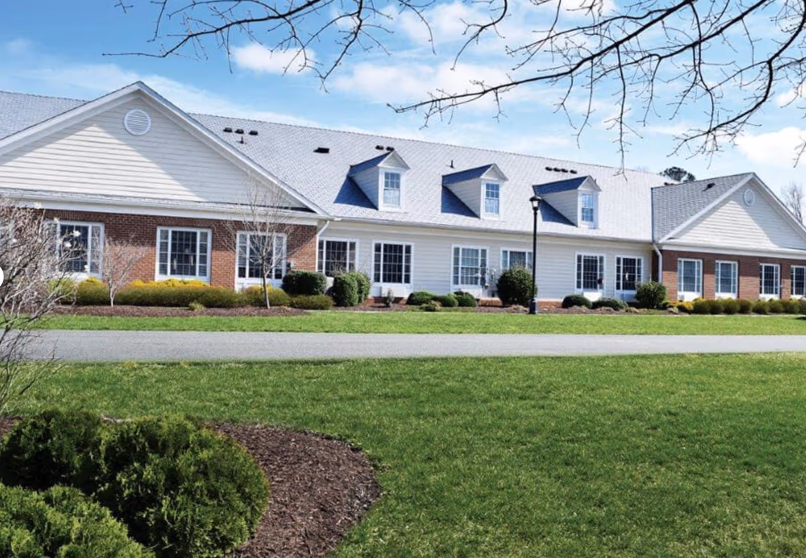 Exterior view of a single-story senior living facility building with a combination of brick and white siding, multiple windows, dormer windows on the roof, a well-maintained lawn, shrubs, and a clear blue sky.