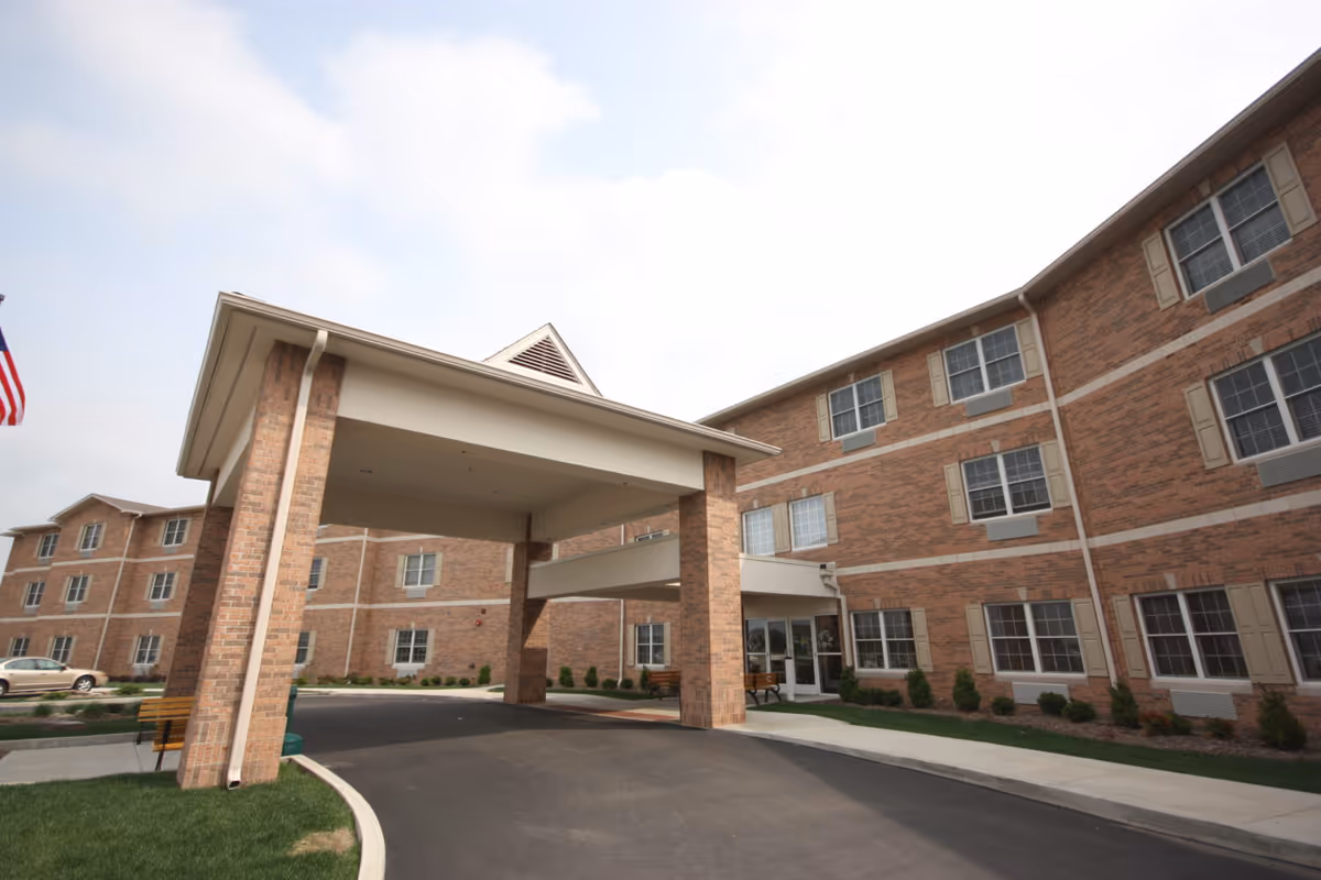Exterior view of a multi-story brick senior living facility with a covered entrance driveway, windows with beige shutters, and a paved driveway surrounded by landscaped greenery.