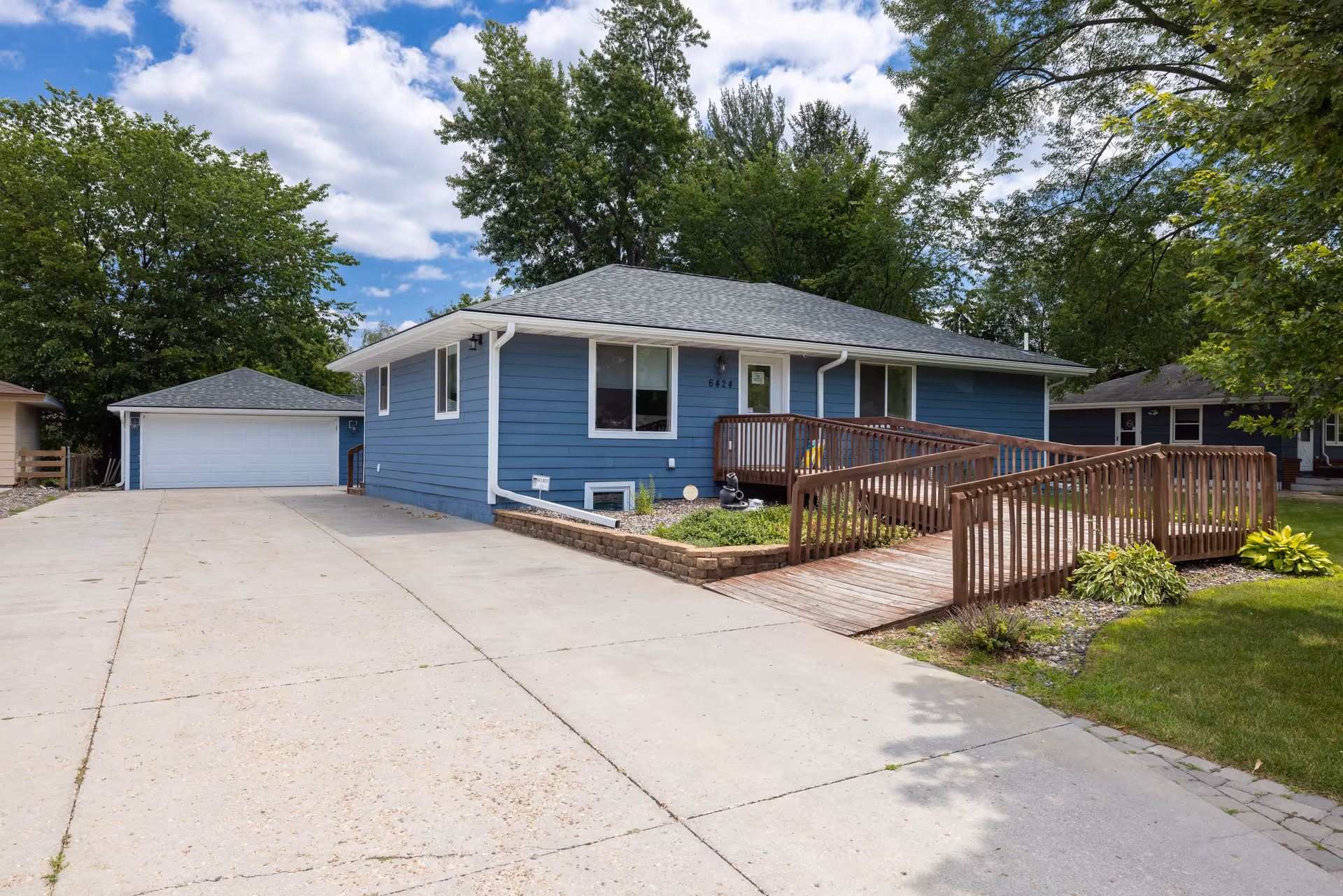 Blue single-story home with wooden ramp, driveway, and surrounding greenery