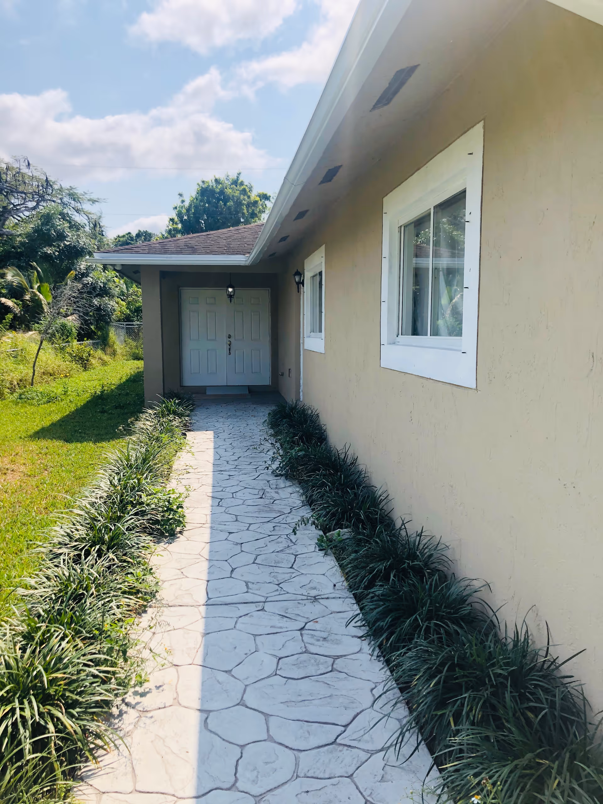 A paved walkway lined with green plants on both sides leading to a beige building with white-framed windows and a double door entrance under a roof overhang. The sky is partly cloudy and there is greenery around the building.