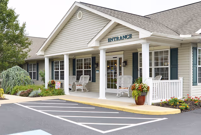 Exterior view of a single-story building with beige siding and a gabled roof. The entrance has a covered porch with white columns, white wicker chairs, potted plants with flowers, and a sign above the door that reads 'ENTRANCE'. There is a parking area with marked spaces in front of the building and landscaping with shrubs and flowers around the porch.