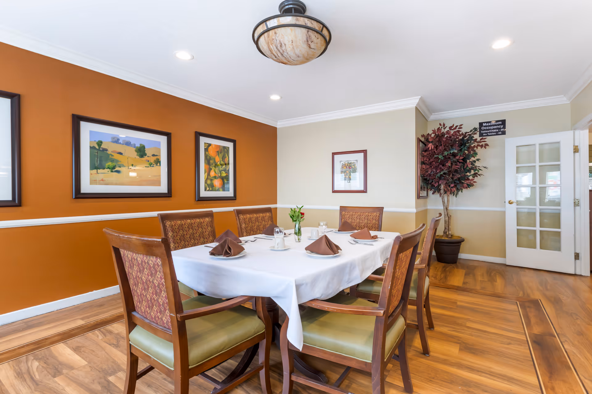 A dining room set with a white-tablecloth table and six wooden chairs, framed artwork on an orange accent wall, and a potted plant by a glass-paneled door.