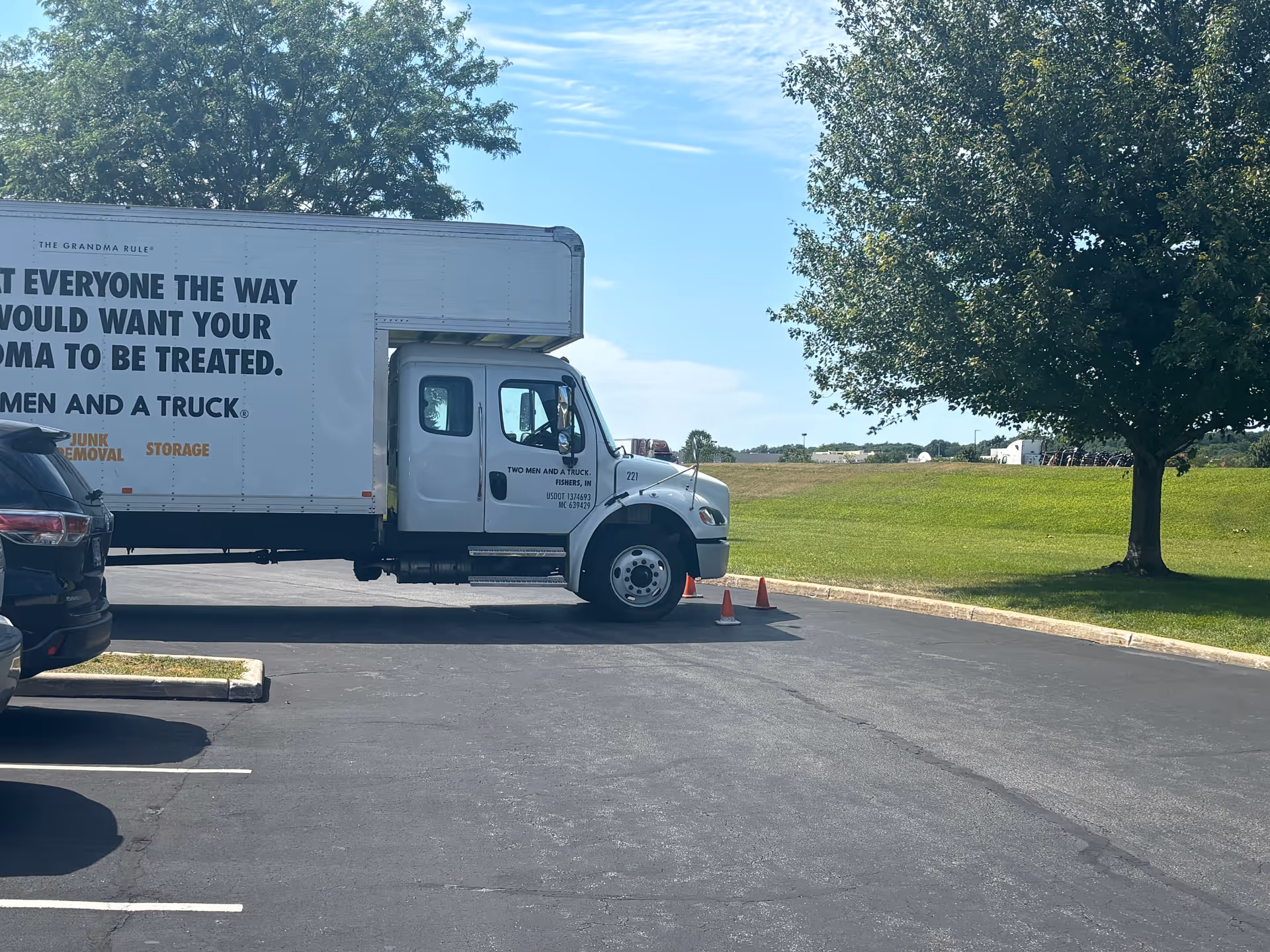 A white box truck parked in a lot beside a grassy field and trees, cones nearby.
