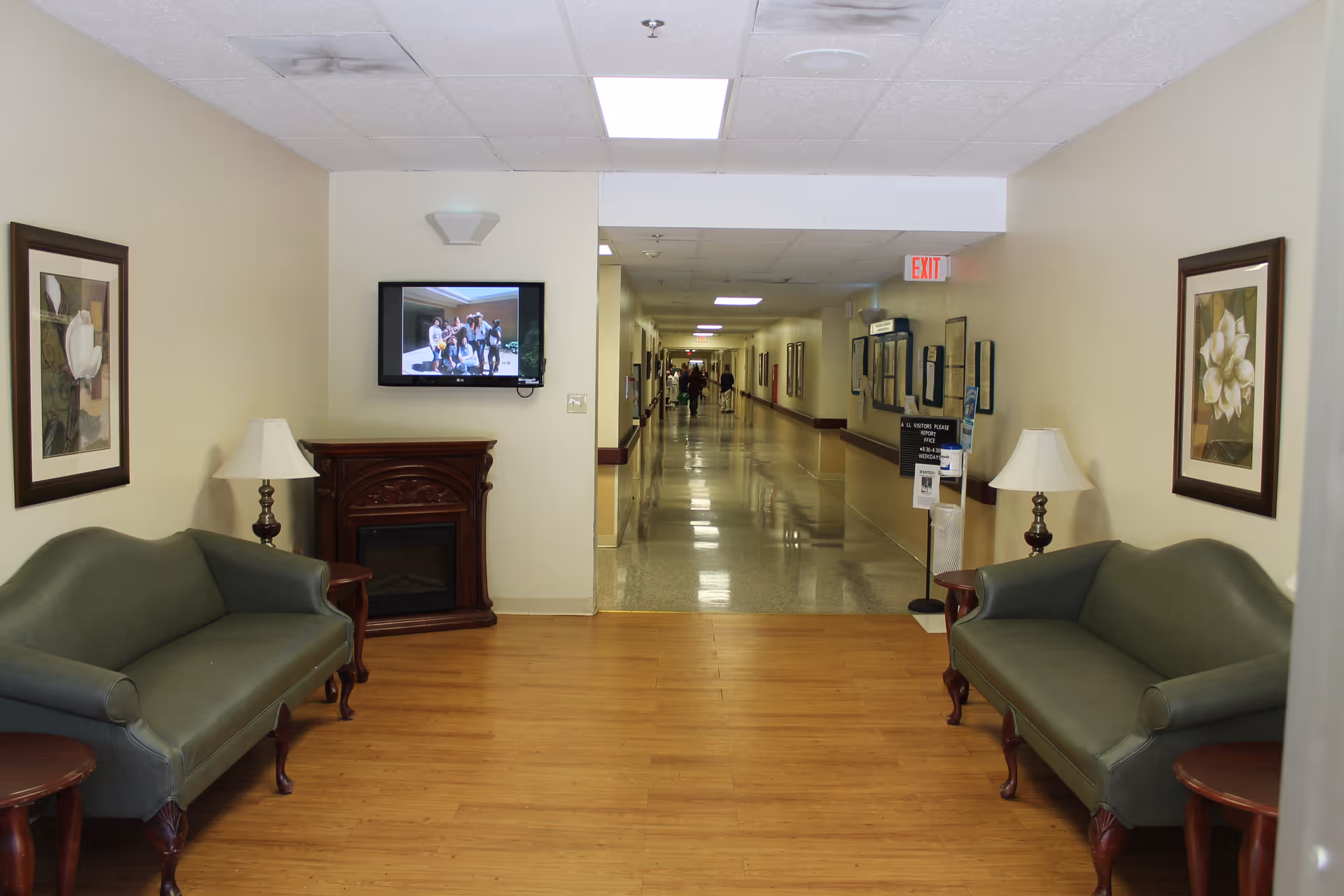 Lobby seating area with two green couches, lamps, framed art, a TV, and a hallway beyond.