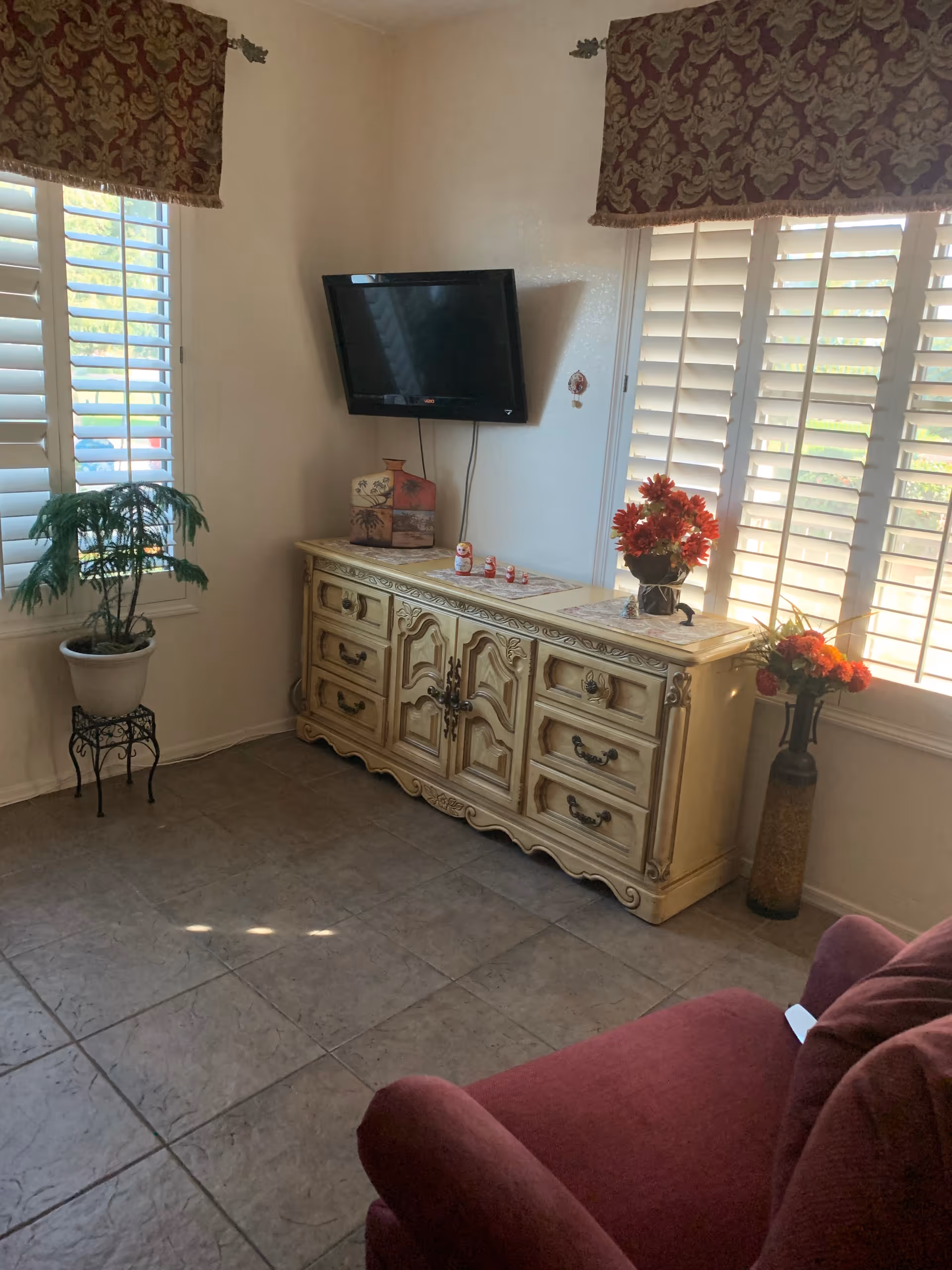 A cream ornate sideboard with a mounted TV, plants, flowers, and shuttered windows.