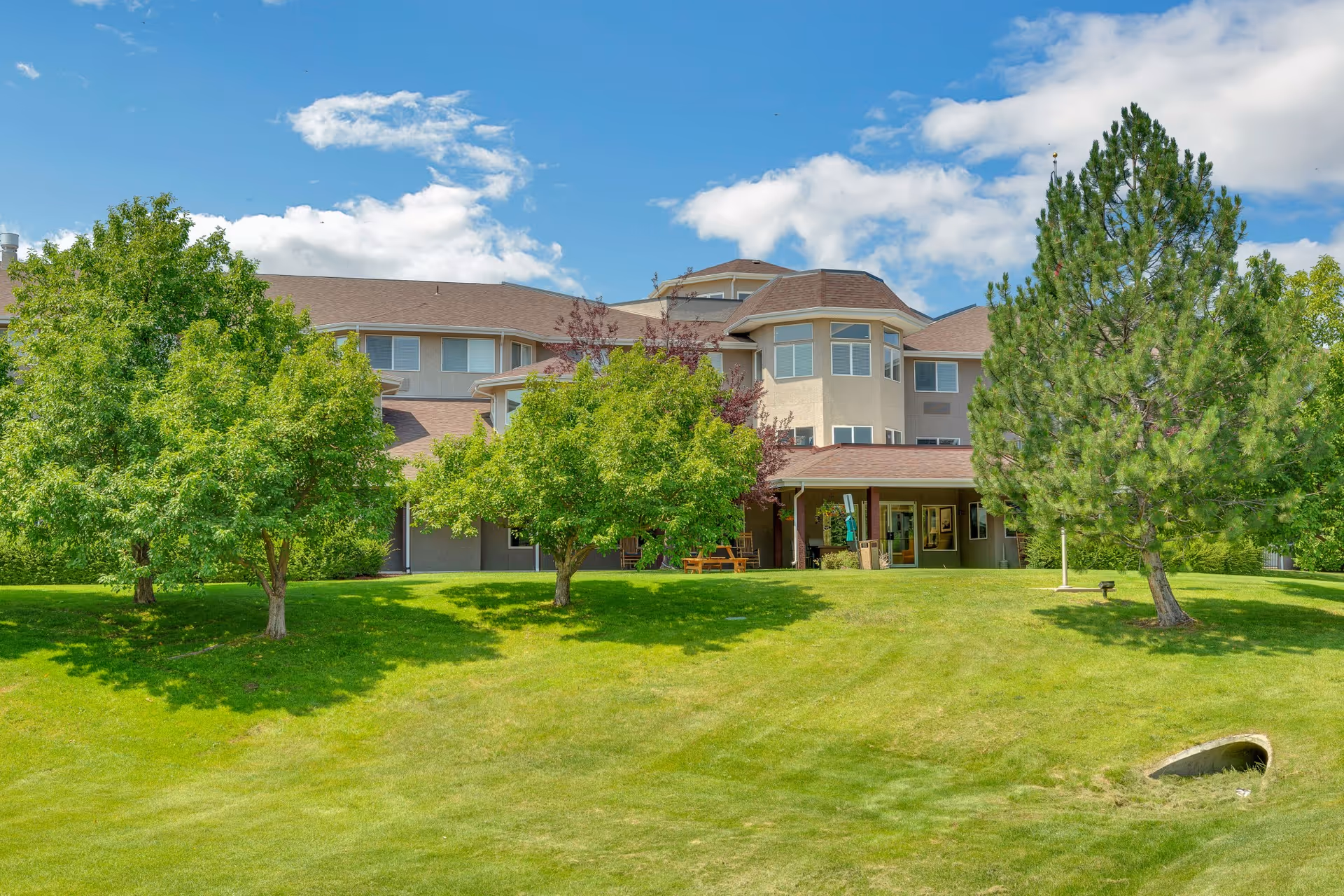 Front exterior of a multi-story senior living building with a grassy lawn, trees, and a blue sky.