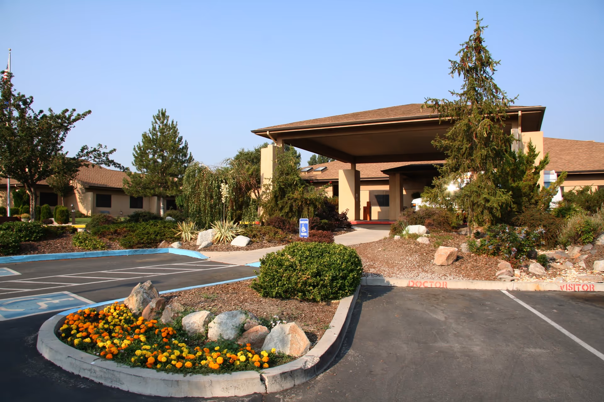 Entrance of a single-story building with a covered drop-off area, surrounded by landscaped bushes, trees, and a flower bed with orange and yellow flowers. There are parking spaces in front, including a handicapped spot and spaces labeled for doctor and visitor parking.