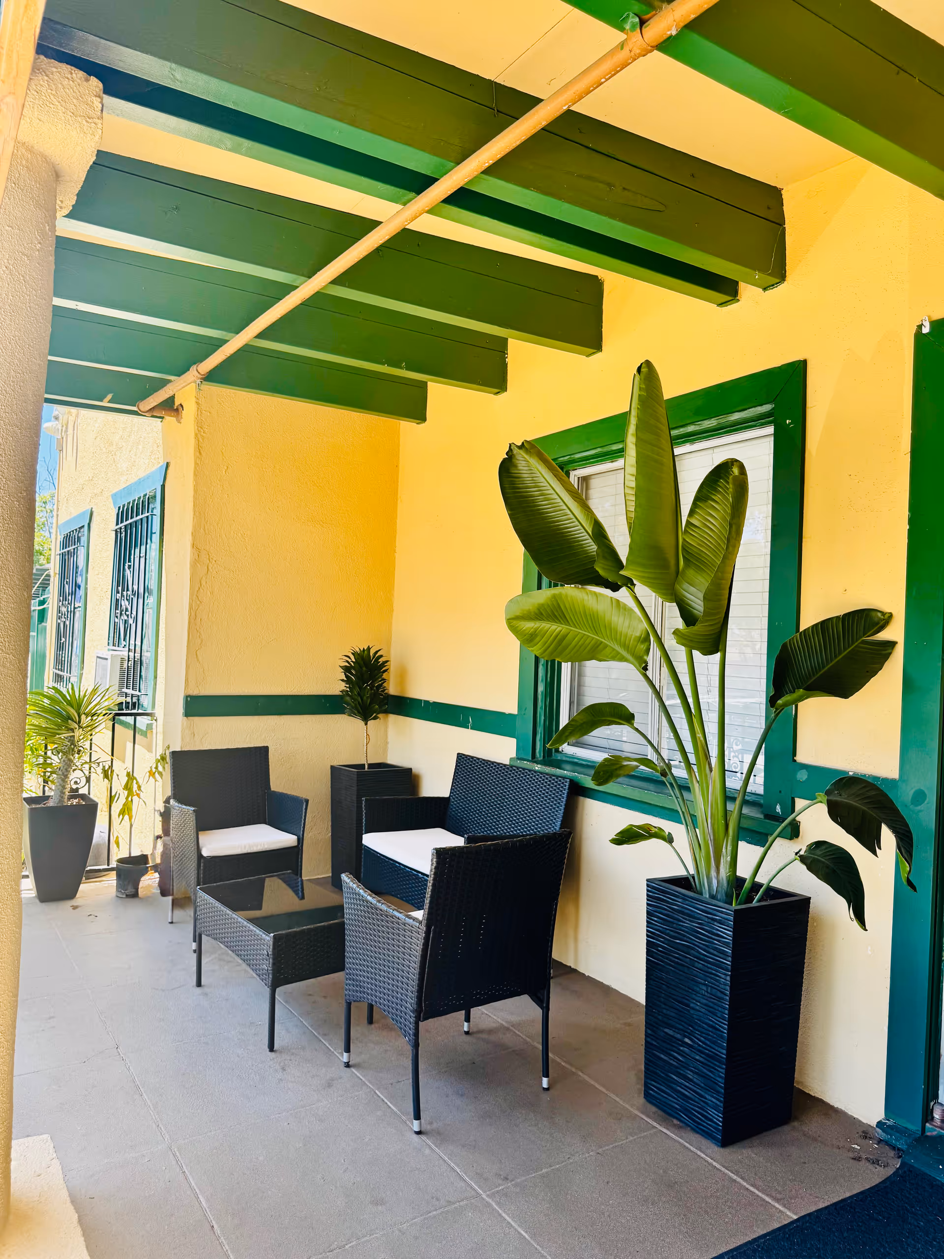 Outdoor veranda with yellow walls, green trim, seating, and potted plants.
