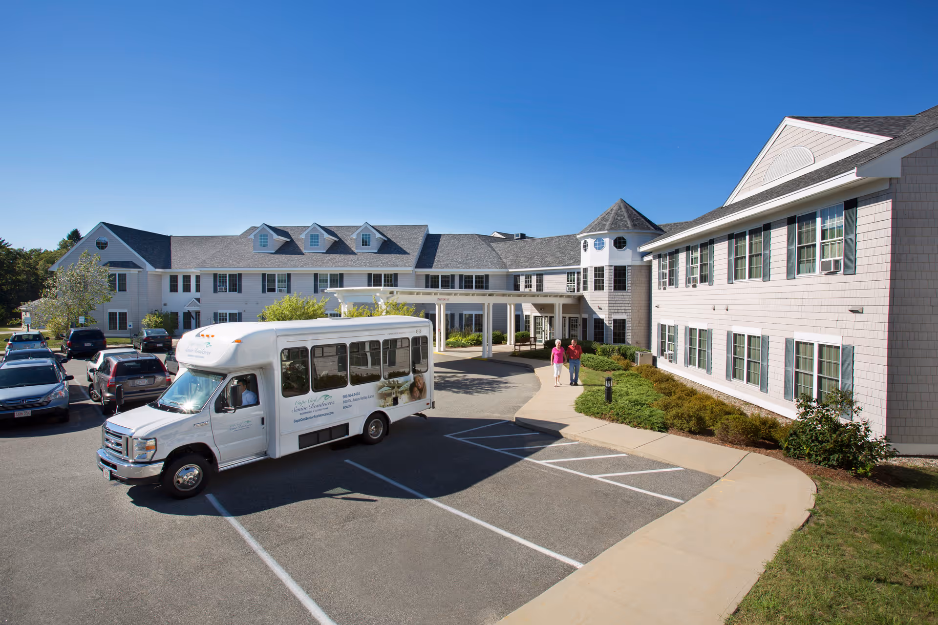 Exterior view of Cape Cod Senior Residences, an Independent and Assisted Living Community, showing a large two-story building with multiple windows, a covered entrance, a parking lot with several cars, a white shuttle bus, and two people walking on the sidewalk near the entrance under a clear blue sky.