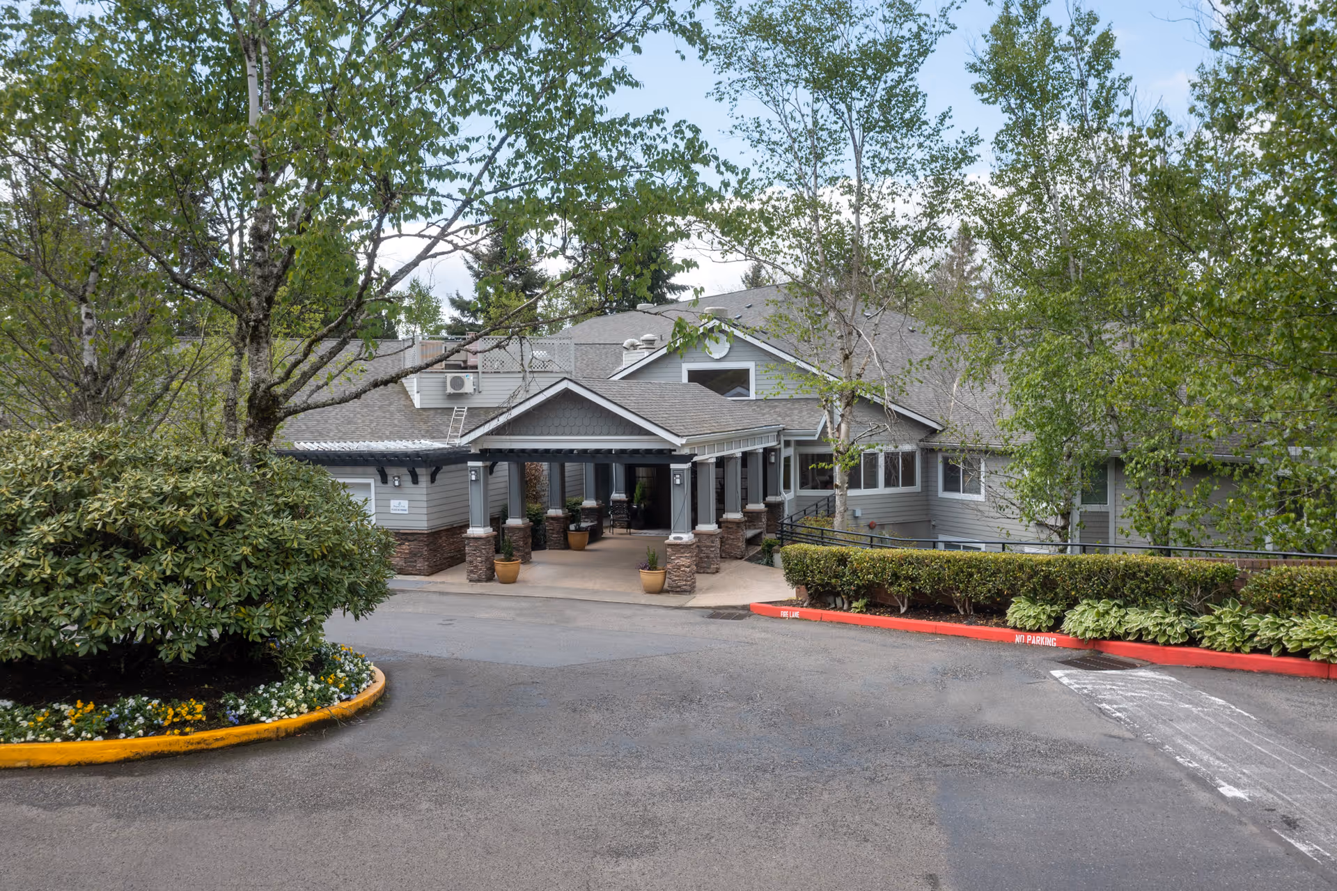 Exterior view of Regency Park senior living facility entrance with a covered driveway, surrounded by trees and landscaped bushes under a partly cloudy sky.