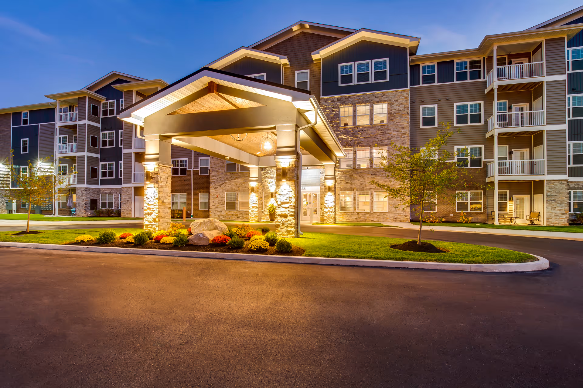 Exterior front entrance of a multi-story apartment building with a lit porte-cochère and landscaped driveway at dusk.