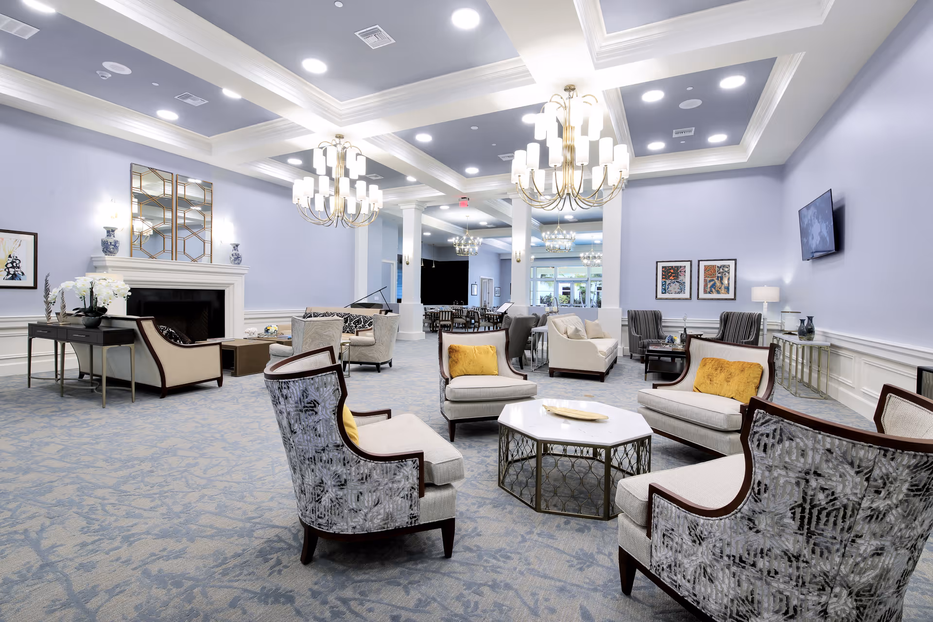 A spacious and elegant senior living common area with multiple seating arrangements including armchairs and sofas around a central coffee table. The room features soft lavender walls, a patterned carpet, large chandeliers, a fireplace with decorative vases, framed artwork, and a wall-mounted television. The area is well-lit with recessed lighting and natural light from windows in the background.