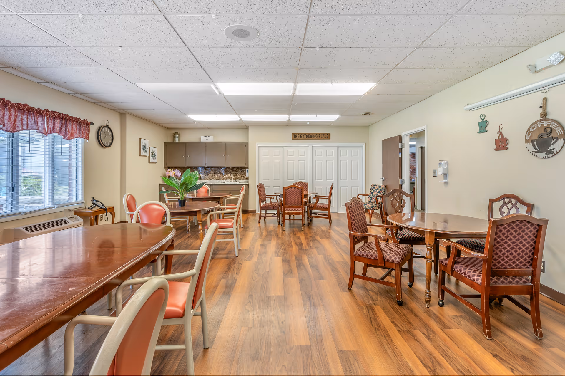 A well-lit communal dining area with several wooden tables and chairs arranged on a wood-patterned floor. The room has a window with red valance curtains on the left, a clock on the wall, and decorative wall hangings including a coffee-themed sign. There is a small kitchenette area with cabinets and a countertop at the back, and a sign above the double doors that reads 'The Gathering Place'.