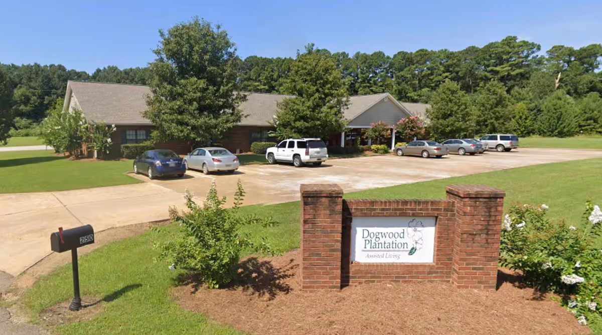Front exterior of a single-story brick assisted living building with a parking lot, several cars, a mailbox, and a brick entrance sign reading "Dogwood Plantation Assisted Living".