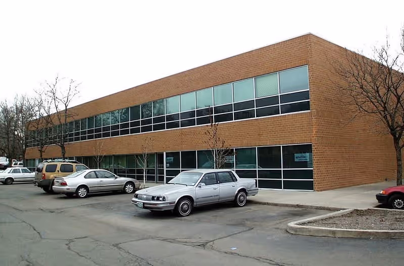 Two-story brick building with large windows and a parking lot in front.